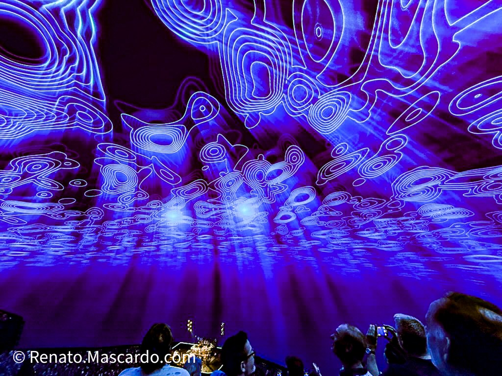 A vibrant light display featuring swirling blue patterns projected onto a ceiling, with concertgoers observing the immersive experience.