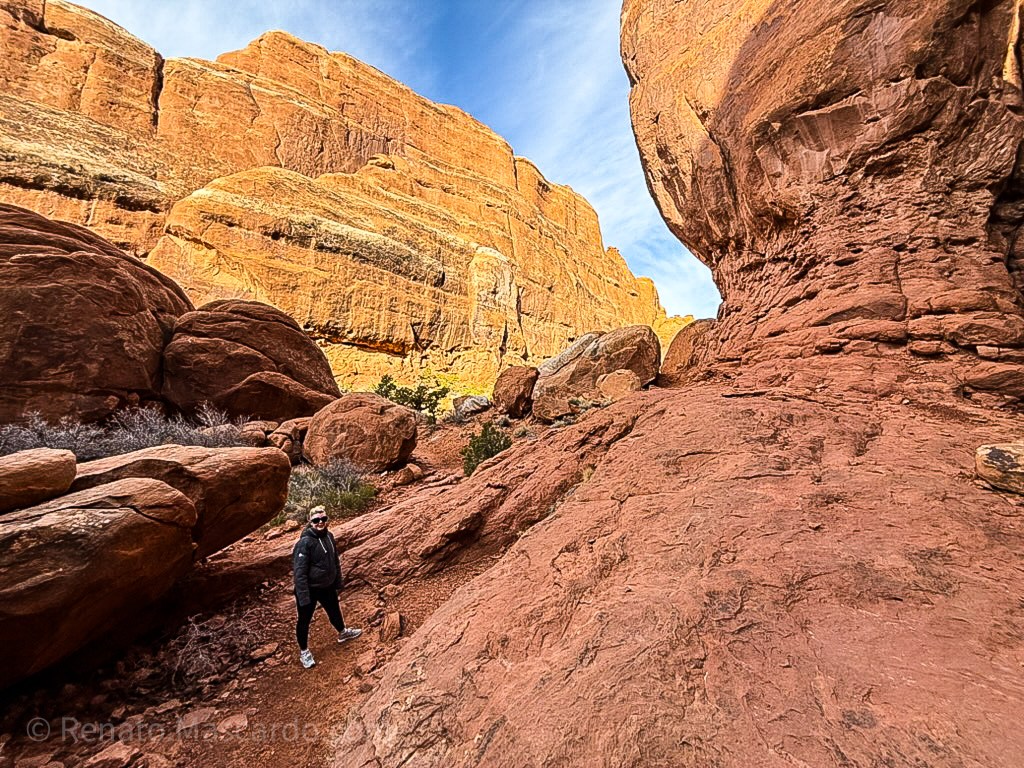 Person standing on a rocky trail surrounded by large red rock formations and a clear blue sky.