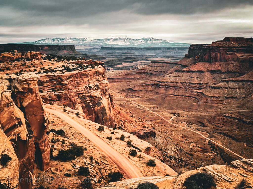 A panoramic view of a rugged landscape with layered rock formations and a winding dirt road cutting through the terrain, under a cloudy sky.