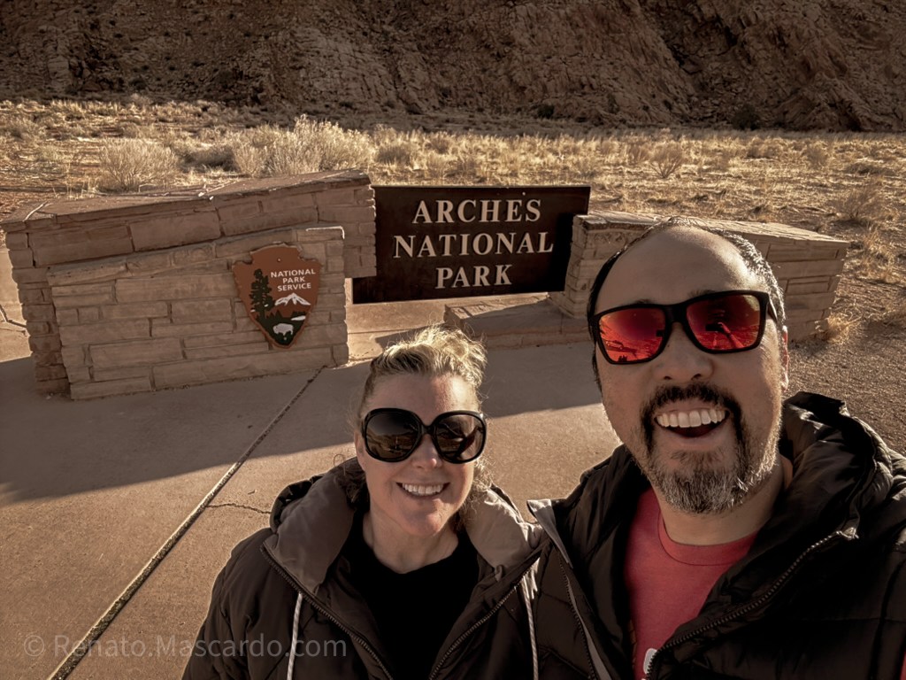 A smiling couple stands in front of the Arches National Park entrance sign, with the park's iconic landscape visible in the background.