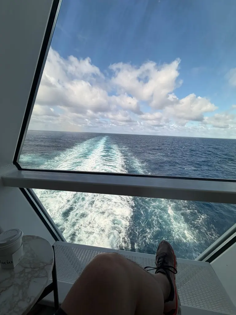 View from a ship cabin showing the ocean and a trail of waves, with a person’s leg and a coffee cup visible in the foreground.