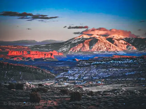 A panoramic view of rugged mountains and cliffs during sunset, showcasing vibrant red and blue hues against the Utah landscape.