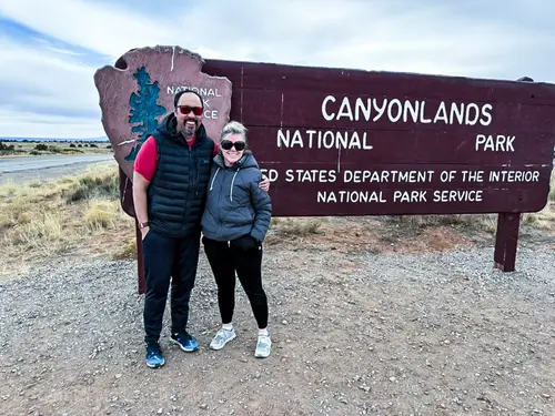 A couple stands in front of the Canyonlands National Park sign, smiling and embracing, with a rugged landscape in the background.